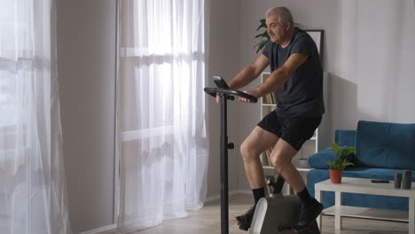 Un-Hombre-Deportivo-De-Mediana-Edad-Está-Entrenando-Con-Una-Bicicleta-Estacionaria-En-Casa,-Tiro-Medio-En-La-Sala-De-Estar,-Un-Estilo-De-Vida-Saludable-En-La-Vejez.